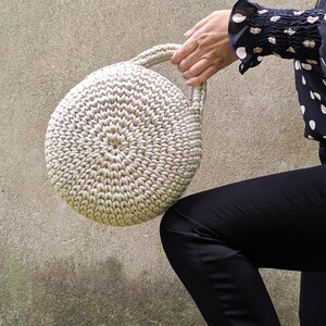 May include: A round, crocheted handbag with a cream-colored design. The bag has a single handle and is held by a person's hand.