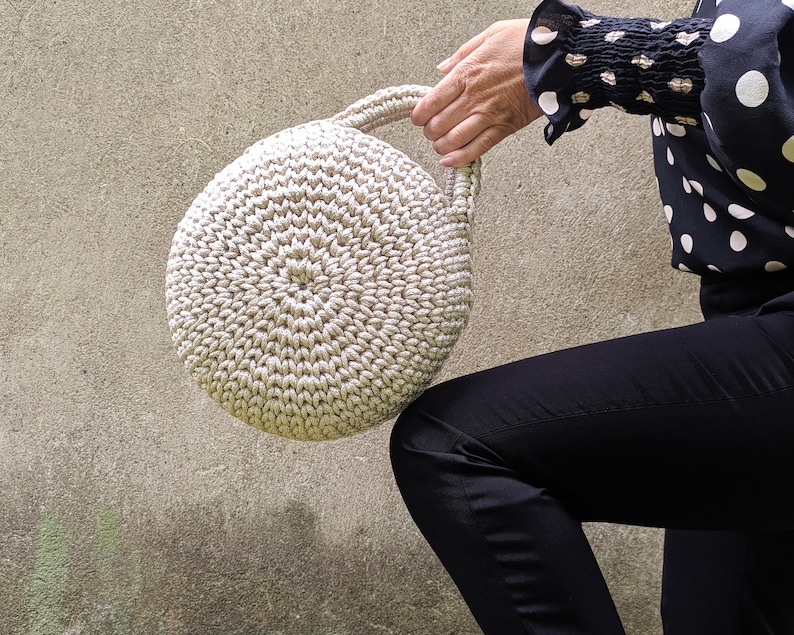 May include: A round, crocheted handbag with a cream-colored design. The bag has a single handle and is held by a person's hand.