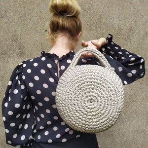May include: A woman wearing a black and white polka dot blouse holds a round, crocheted, cream-colored handbag.