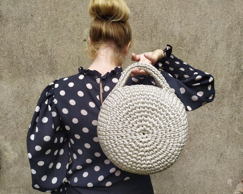 May include: A woman wearing a black and white polka dot blouse holds a round, crocheted, cream-colored handbag.