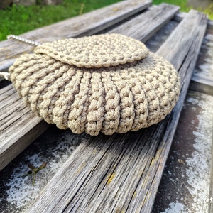 Pu&ograve; includere: Una borsa a mano in crochet beige con chiusura a lembo e una tracolla a catena argentata. La borsa &egrave; appoggiata su una superficie di legno.