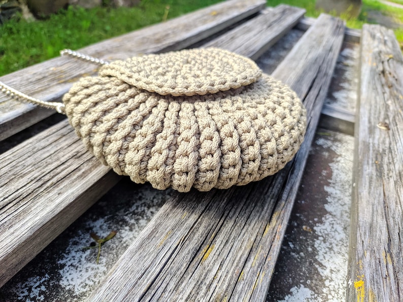 Pu&ograve; includere: Una borsa a mano in crochet beige con chiusura a lembo e una tracolla a catena argentata. La borsa &egrave; appoggiata su una superficie di legno.