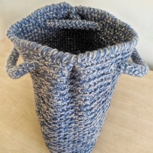 May include: A blue and white knitted basket with two small handles. The basket is cylindrical with a slightly flared opening. The knit pattern is visible, creating a textured surface. The interior is dark, contrasting with the lighter exterior.