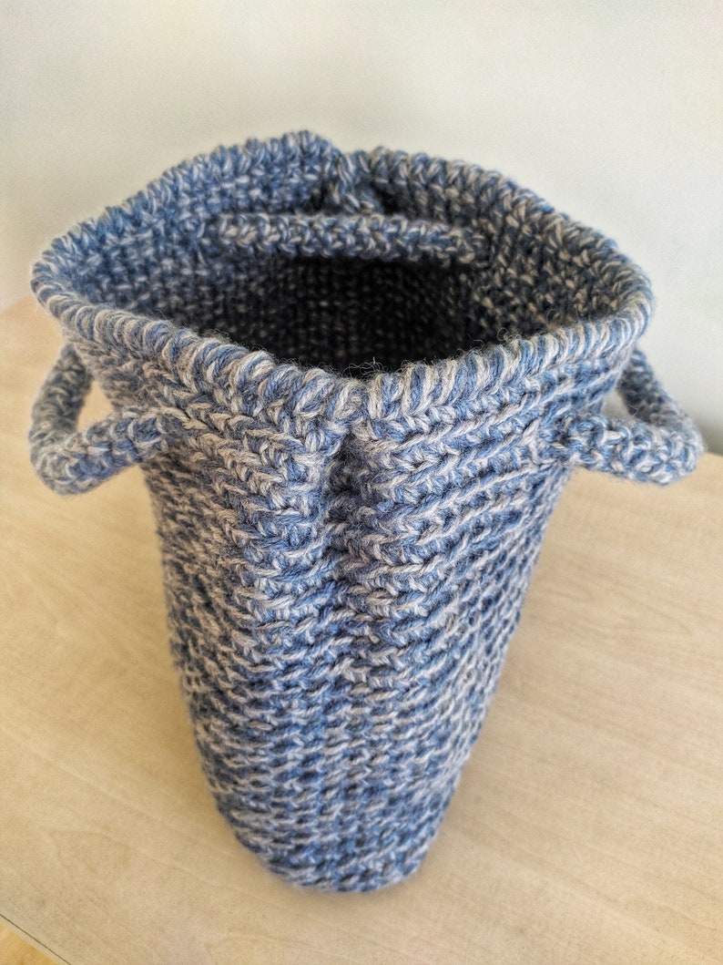 May include: A blue and white knitted basket with two small handles. The basket is cylindrical with a slightly flared opening. The knit pattern is visible, creating a textured surface. The interior is dark, contrasting with the lighter exterior.