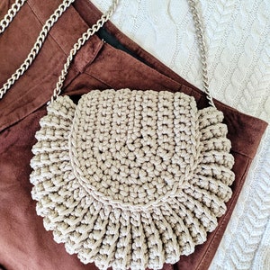 Pu&ograve; includere: Una borsa a mezza luna in crochet beige con una tracolla a catena argentata. La borsa &egrave; appoggiata su una superficie in camoscio marrone.