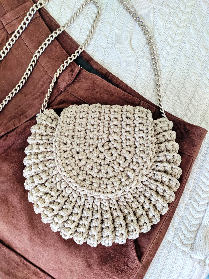 Pu&ograve; includere: Una borsa a mezza luna in crochet beige con una tracolla a catena argentata. La borsa &egrave; appoggiata su una superficie in camoscio marrone.