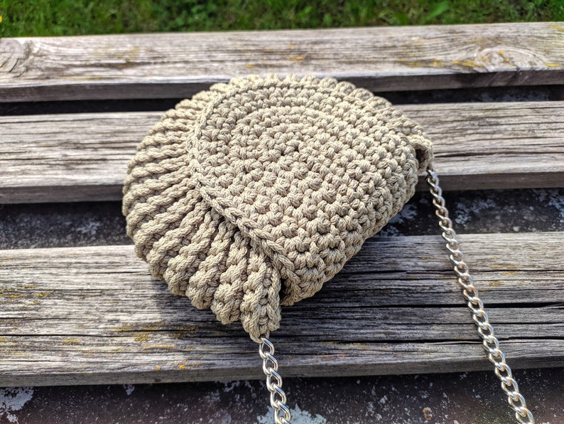 Pu&ograve; includere: Una borsa in crochet beige con una tracolla a catena argentata. La borsa &egrave; appoggiata su una superficie di legno.