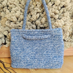 May include: A blue and white woven tote bag with two handles. The bag has a rectangular shape and a textured appearance. The handles are also woven and are a lighter shade of blue. The bag is sitting on a wooden surface.