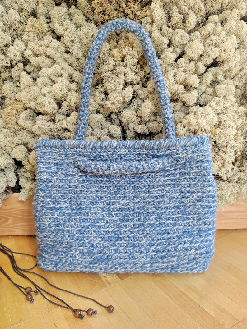 May include: A blue and white woven tote bag with two handles. The bag has a rectangular shape and a textured appearance. The handles are also woven and are a lighter shade of blue. The bag is sitting on a wooden surface.
