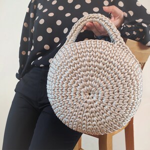 May include: A round, crocheted handbag with a beige and silver color scheme. The bag has a single handle and a circular shape.