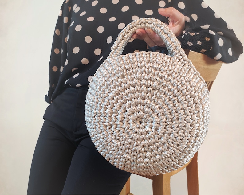 May include: A round, crocheted handbag with a beige and silver color scheme. The bag has a single handle and a circular shape.