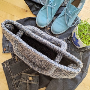 May include: A blue and white crocheted tote bag sits atop a denim jacket and jeans. Light blue suede lace-up shoes are also visible. A small white and blue floral planter with greenery is in the background. The items are arranged on a light wood surface.
