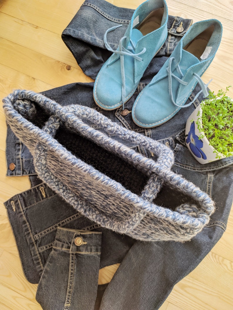 May include: A blue and white crocheted tote bag sits atop a denim jacket and jeans. Light blue suede lace-up shoes are also visible. A small white and blue floral planter with greenery is in the background. The items are arranged on a light wood surface.