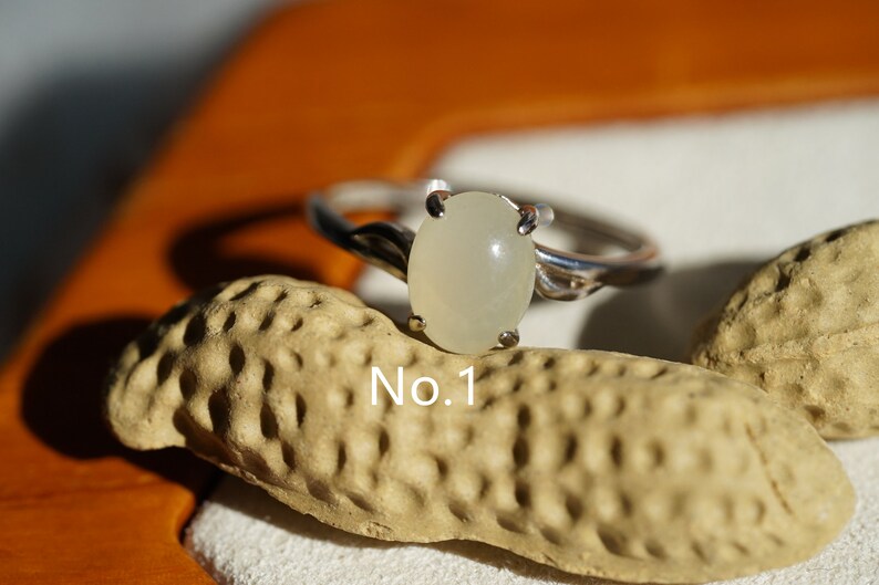 May include: A silver ring with a white oval gemstone. The ring is set on a textured surface with the text "No. 1" visible.