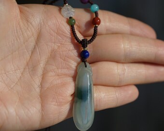 Puede incluir: Un collar con un colgante de jade verde y blanco, un cordón marrón y cuentas de colores. Las cuentas son verdes, azules, rojas y blancas.