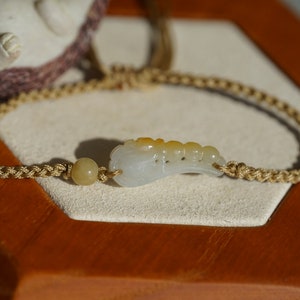 Peut inclure: Un bracelet en jade blanc avec un motif sculpté, comprenant une perle brun clair et un cordon tressé.