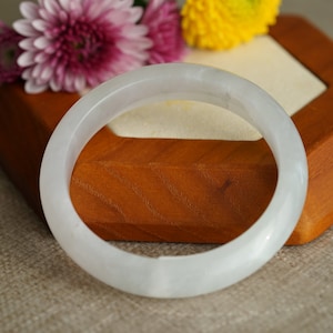 May include: A white jade bangle bracelet on a wooden display stand. The bangle is smooth and polished, with a subtle sheen.
