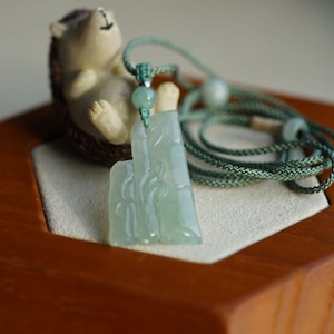 May include: A jade pendant necklace with a carved bamboo design. The pendant is light green and has a smooth, polished finish. The necklace is made of a green cord and has a small green bead.