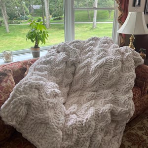 May include: A large, off-white, chunky knit blanket with a cable-knit design. The blanket is draped over a patterned sofa, creating a cozy and inviting aesthetic. The soft texture and neutral color make it a versatile home decor item.
