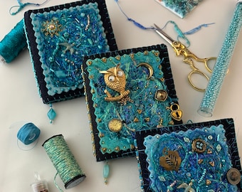 May include: Three small blue and black fabric covered journals with gold embellishments and beads. The journals are decorated with an owl, a heart, and a bird. There are also various beads and other embellishments scattered around the journals.