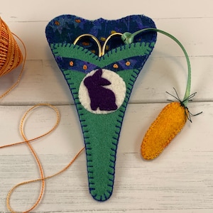 May include: A green felt scissor holder with a purple bunny applique and a carrot-shaped loop. The holder is decorated with blue stitching and orange dots.