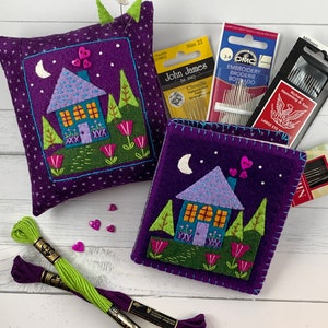 May include: Purple felt pincushion and needle book with a whimsical house design, featuring a moon, stars, trees, and flowers. The pincushion has a button in the shape of a heart. The needle book has the text "John James Est. 1843 Chenille" on the cover.
