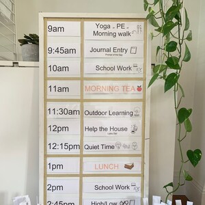 May include: A white framed daily schedule board with labeled time slots. Activities include "Yoga or PE", "Journal Entry", "School Work", "Morning Tea", "Outdoor Learning", "Help the House", "Quiet Time", "Lunch", and "Pack Away the Day".