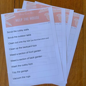 May include: Several white paper sheets with the text "HELP THE HOUSE" and a list of chores. The top of the sheets have a peach-colored banner with illustrations of a pencil, ruler, and other school supplies. The sheets are stacked on a wooden surface.