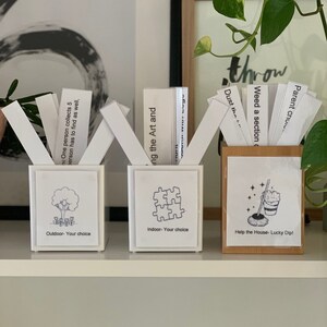 May include: Three decorative boxes with white cards. The first box has a tree graphic and text "Outdoor - Your choice". The second box has a puzzle graphic and text "Indoor - Your choice". The third box is wooden with a mop graphic and text "Help the House - Lucky Dip!"