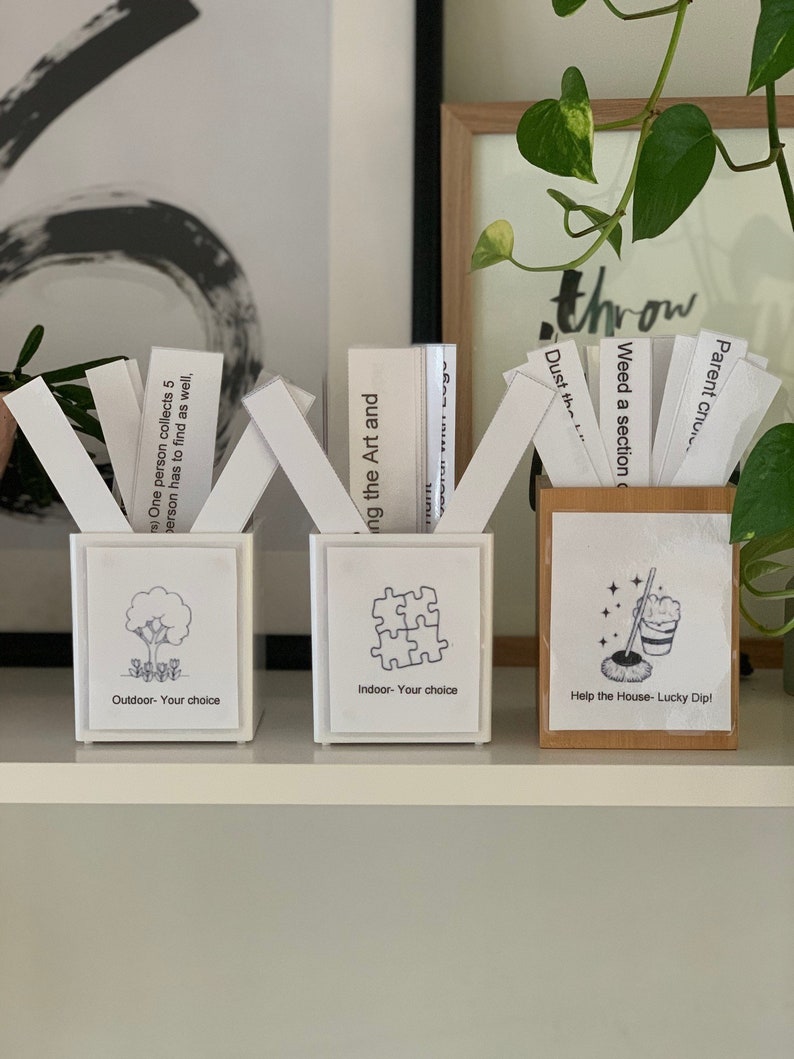 May include: Three decorative boxes with white cards. The first box has a tree graphic and text "Outdoor - Your choice". The second box has a puzzle graphic and text "Indoor - Your choice". The third box is wooden with a mop graphic and text "Help the House - Lucky Dip!"
