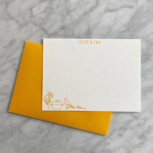 Puede incluir: Una tarjeta de papelería rectangular blanca con el texto dorado JOCIE & TIM en la parte superior y dibujos lineales dorados de un lirio, una olla, una paloma y un batidor en la esquina inferior izquierda, junto a un sobre amarillo mostaza.