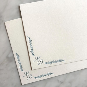 Peut inclure: Deux feuilles de papeterie blanc cassé avec des motifs botaniques d'angle bleu sarcelle et les initiales "MD". Les feuilles encadrent les initiales. Le papier a une surface texturée, sur fond marbré.