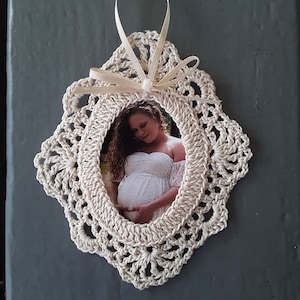 Pode incluir: Um quadro de crochê branco com uma fita de cetim branca e uma pérola. Uma foto de uma mulher grávida está dentro do quadro.