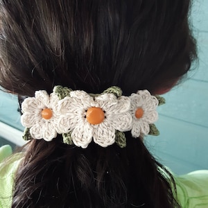 May include: A white crocheted hair tie with three daisy flowers and orange centres. The flowers are connected by green crocheted leaves.