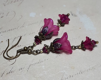 Puede incluir: Un par de pendientes de flores rosas con detalles en bronce. Los pendientes tienen un encanto de flor colgante con una cuenta de cristal y un cierre de gancho.