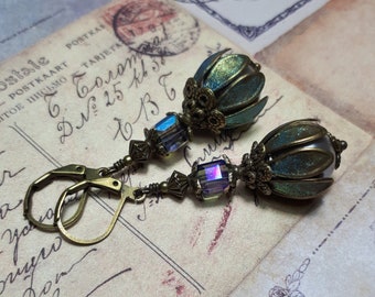 Puede incluir: Un par de pendientes con cuentas de color azul y verde iridiscente en forma de lágrima, acentuadas con metal dorado y detalles de cristal.