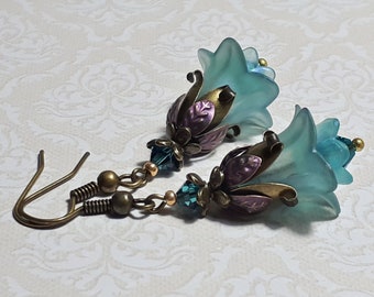 May include: A pair of earrings featuring blue glass flower charms with bronze and purple accents. The earrings have a bronze chain and hook.