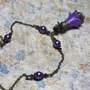 Vintage Style Deadly nightshade inspired necklace that has a purple flower drop with topaz yellow crystals and antique floral bead caps. It has a bronze chain that has purple pearls and crystals spaced along it. It has a whimsigoth style to it.