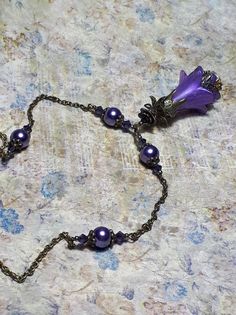 Vintage Style Deadly nightshade inspired necklace that has a purple flower drop with topaz yellow crystals and antique floral bead caps. It has a bronze chain that has purple pearls and crystals spaced along it. It has a whimsigoth style to it.