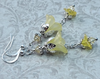 May include: A pair of delicate earrings featuring pale yellow flower-shaped pendants. The earrings have silver-colored metal accents and small, clear beads. The hooks are silver-colored. The earrings are displayed on a patterned background.
