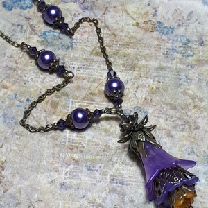 Vintage Style Deadly nightshade inspired necklace that has a purple flower drop with topaz yellow crystals and antique floral bead caps. It has a bronze chain that has purple pearls and crystals spaced along it. It has a whimsigoth style to it.