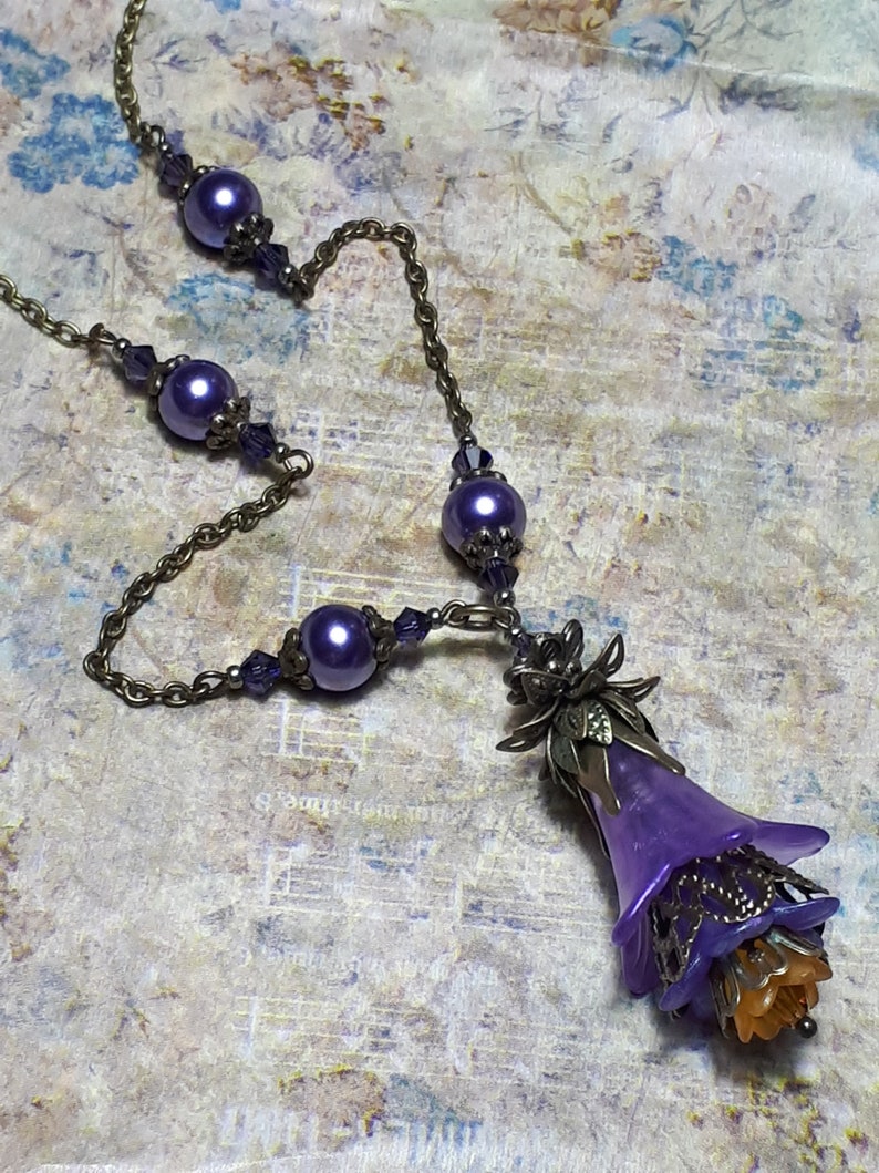 Vintage Style Deadly nightshade inspired necklace that has a purple flower drop with topaz yellow crystals and antique floral bead caps. It has a bronze chain that has purple pearls and crystals spaced along it. It has a whimsigoth style to it.