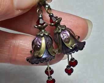 May include: A pair of dangle earrings with a bell-shaped design. The bells are made of metal with a purple and gold finish. The earrings are accented with red glass beads.
