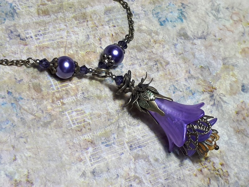 Vintage Style Deadly nightshade inspired necklace that has a purple flower drop with topaz yellow crystals and antique floral bead caps. It has a bronze chain that has purple pearls and crystals spaced along it. It has a whimsigoth style to it.