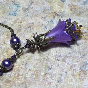 Vintage Style Deadly nightshade inspired necklace that has a purple flower drop with topaz yellow crystals and antique floral bead caps. It has a bronze chain that has purple pearls and crystals spaced along it. It has a whimsigoth style to it.