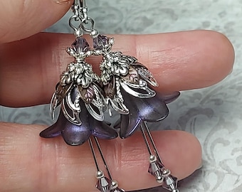 Puede incluir: Pendientes colgantes con colgantes en forma de flor púrpura. Los pendientes tienen detalles metálicos plateados y pequeñas cuentas colgantes. Los pétalos de la flor son de color púrpura intenso y el diseño es intrincado y detallado.