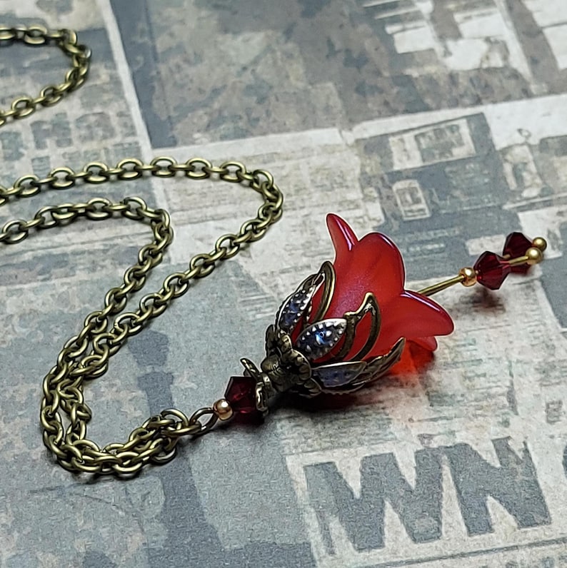 A red vintage style Lucite flower pendant necklace with a bronze filigree design and Siam red crystal beads that hang and sway from under the flowers. The necklace has a bronze tone chain. Fairycore style jewelry gift. Whimsical Necklace.