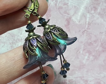 Puede incluir: Pendientes colgantes con diseño floral. Los pendientes presentan una flor translúcida azul en forma de campana con detalles en verde y morado. Están adornados con cristales azul oscuro y detalles metálicos dorados.