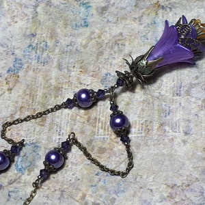 Vintage Style Deadly nightshade inspired necklace that has a purple flower drop with topaz yellow crystals and antique floral bead caps. It has a bronze chain that has purple pearls and crystals spaced along it. It has a whimsigoth style to it.