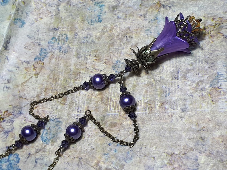 Vintage Style Deadly nightshade inspired necklace that has a purple flower drop with topaz yellow crystals and antique floral bead caps. It has a bronze chain that has purple pearls and crystals spaced along it. It has a whimsigoth style to it.